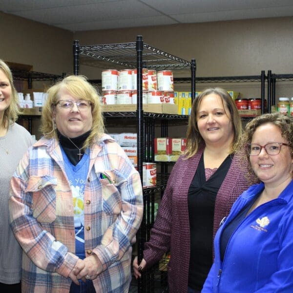Group of four women standing together in pantry with shelves stacked with parishable goods.