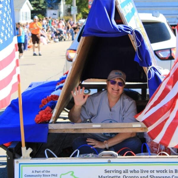 Happy women sitting inside parade float with American flags surrounding her and a large crowd in the background.