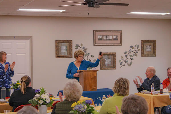 Woman standing at podium making speech while people sit, listen and clap.