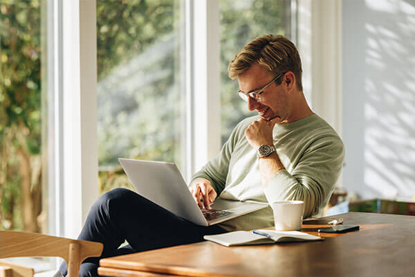 Pensive man sitting at table with laptop resting in lap with notebook, coffee cup and phone on surface.