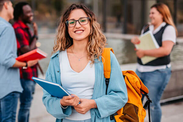 Female student standing in front of other colleagues holding book in her hands and backpack on shoulder.