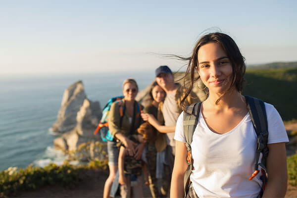 Girl standing in foreground smiling with backpack on back while friends huddle behind her while they all stand on side of mountain overlooking the ocean.