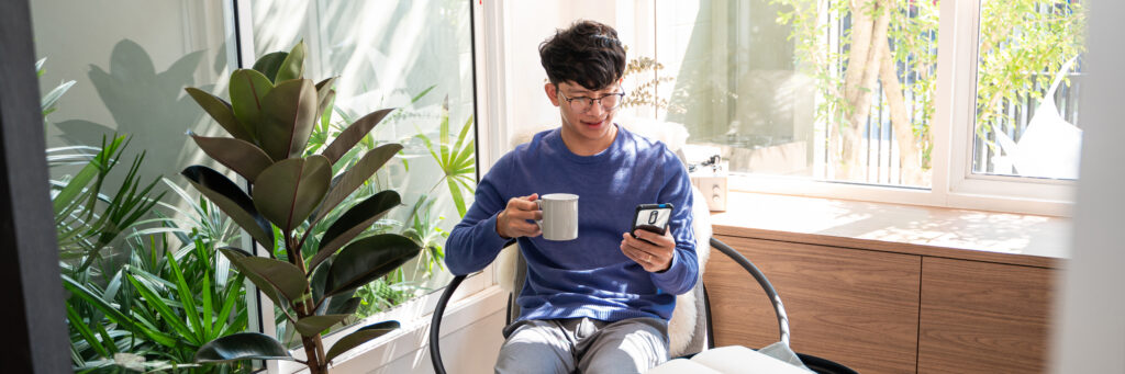 Handsome Asian teenager man wearing casual clothes in glasses using smart phone browsing Internet on mobile phone device at the living room.